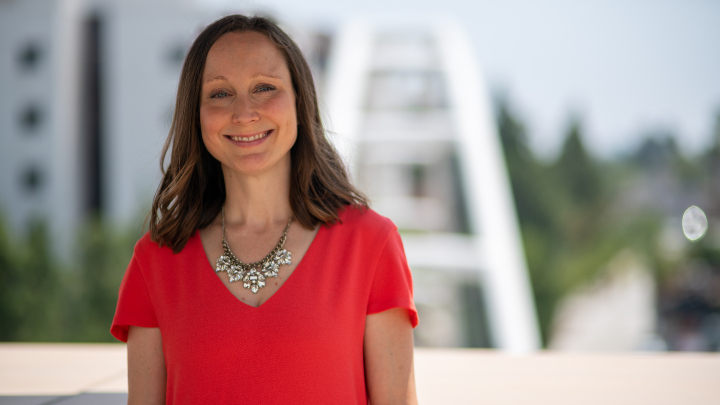 A woman in a red shirt smiles as she stands in front of a bridge.