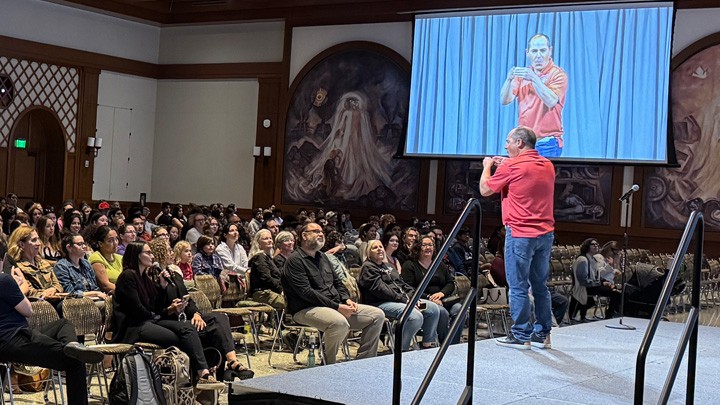 A man in a red shirt signs to a large group of people in a ballroom.