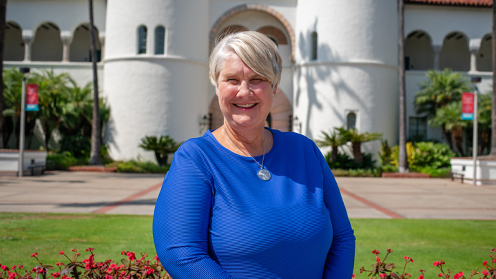 A woman in blue stands in front of a white building with Spanish archways.
