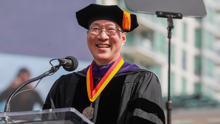 A man in black commencement regalia speaks at a podium.