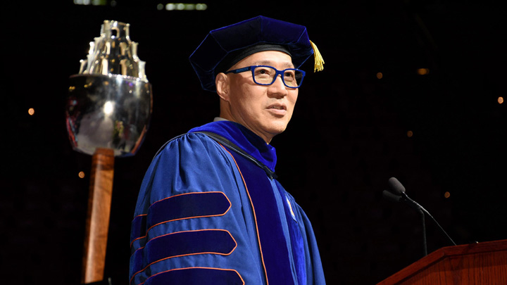 A man in blue doctoral attire stands on stage at commencement.