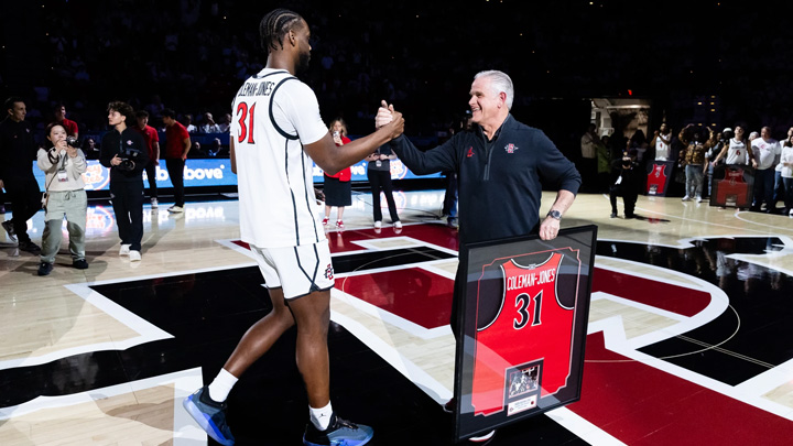 A basketball player shakes hands with a coach holding a framed jersey.
