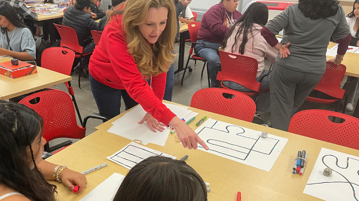 A woman in red points to science activities on a table while two young students look on.
