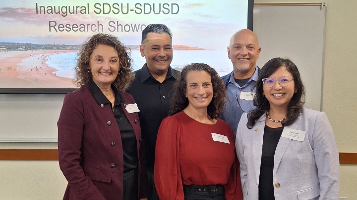 Five people smile in front of the projected words "Inaugural SDSU-SDUSD Research Showcase"