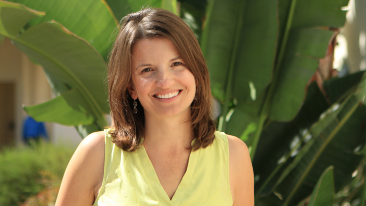 A woman in yellow smiles in front of greenery.