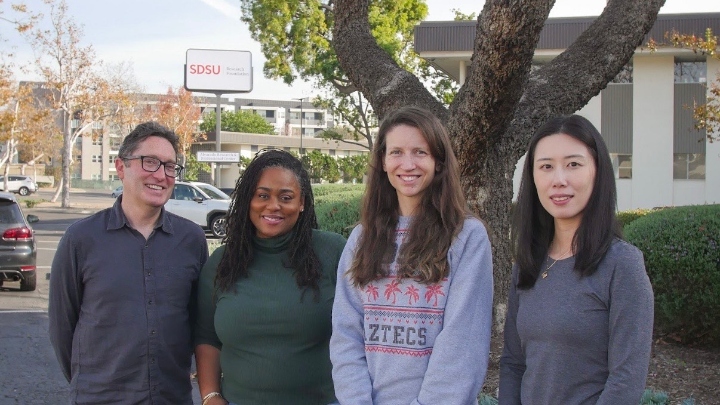 Four people stand in front of trees and a white building.