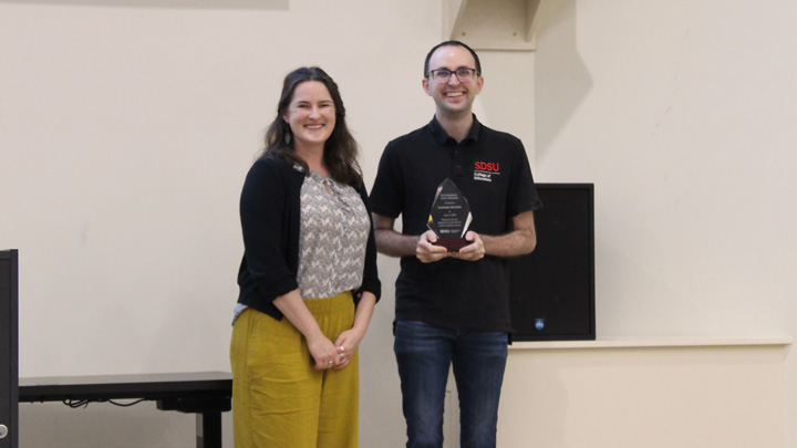 A man in a black polo holds an award while standing next to a woman in a black cardigan.