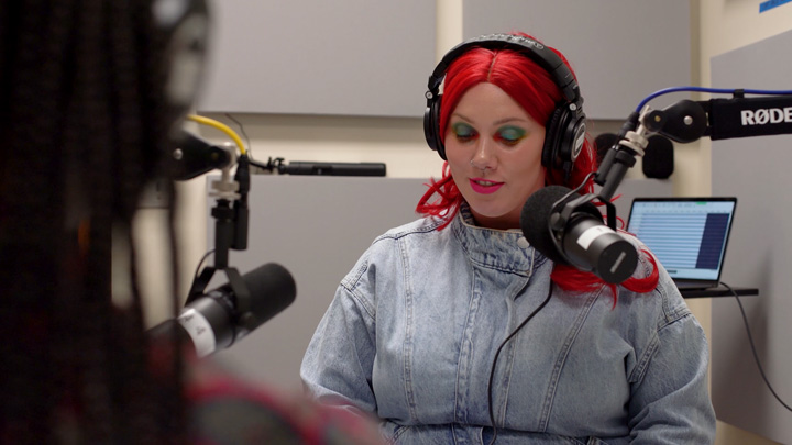 A woman in a denim jacket speaks into a podcasting microphone.