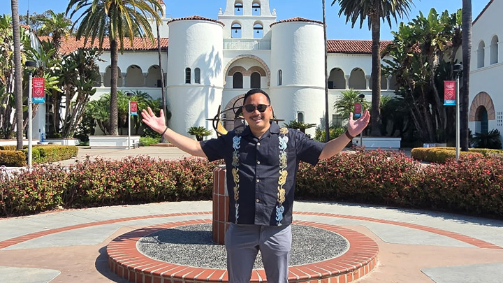 A man in a black shirt stands in front of a white building and palm trees.